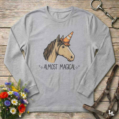 A heather gray long-sleeve t-shirt with a graphic of a horse head wearing a cowboy hat and the text 'ALMOST MAGICAL' underneath, displayed in a flat-lay on a rustic wooden background with flowers and leather tack.