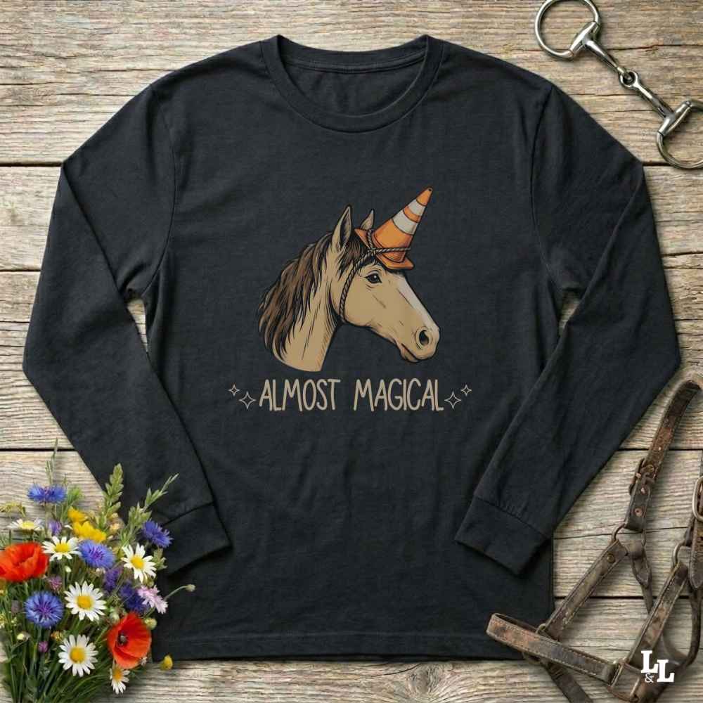 A black long-sleeve t-shirt with a graphic of a unicorn wearing an orange and white traffic cone horn, with the text 'ALMOST MAGICAL' printed below. The shirt is displayed in a flat-lay arrangement on a rustic wooden background, surrounded by decorative props including a bouquet of flowers, a horse bridle, and a metal tack clip.