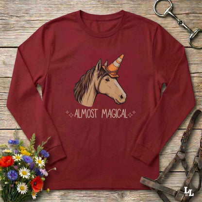 A long-sleeve crewneck t-shirt in maroon red, laid flat on a rustic wooden surface. The shirt features a graphic of a unicorn with a small orange cone hat and the text 'ALMOST MAGICAL' printed below.