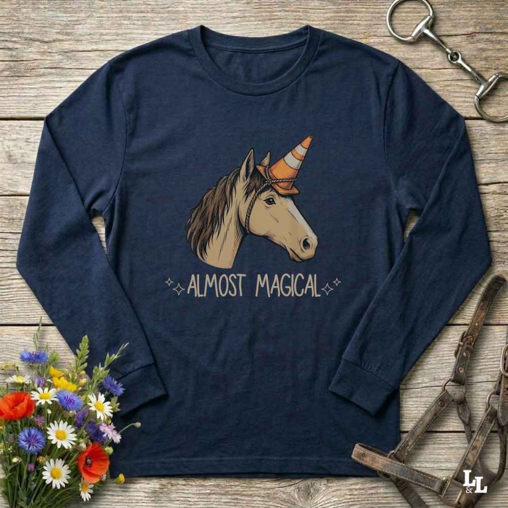 A navy blue long-sleeve t-shirt with a graphic of a unicorn wearing an orange and white striped horn, with the text 'ALMOST MAGICAL' printed below. The shirt is displayed in a flat-lay arrangement on a rustic wooden background, surrounded by decorative elements including a bouquet of flowers, a horse bridle, and a horse halter.