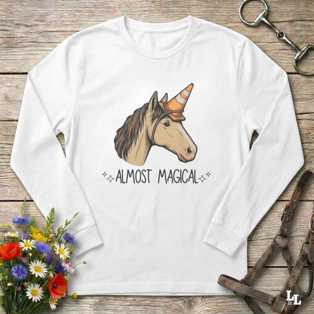 A white long-sleeve t-shirt with a graphic of a unicorn wearing an orange and black horn, with the text 'ALMOST MAGICAL' printed below. The shirt is displayed flat on a rustic wooden background with decorative floral and equestrian props.