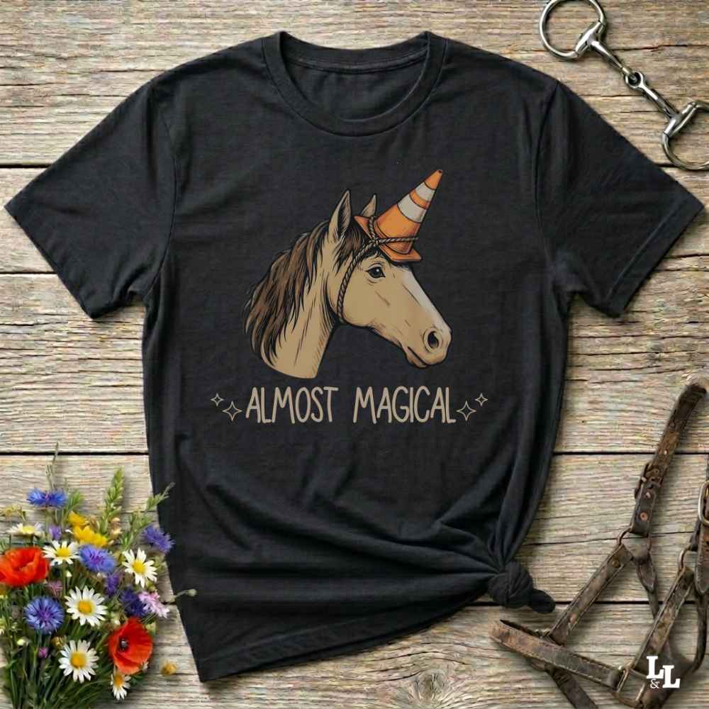 A black short-sleeve graphic t-shirt laid flat on a rustic wooden surface, featuring an illustration of a horse head with a traffic cone horn and the text 'ALMOST MAGICAL'. The shirt is styled with equestrian accessories (bridle) and wildflowers.