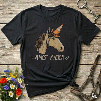 A black short-sleeve graphic t-shirt laid flat on a rustic wooden surface, featuring an illustration of a horse head with a traffic cone horn and the text 'ALMOST MAGICAL'. The shirt is styled with equestrian accessories (bridle) and wildflowers.