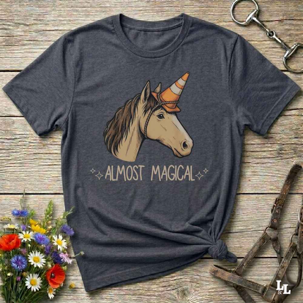 A dark heather gray graphic t-shirt laid flat on a rustic wooden surface. The shirt features a graphic of a white and brown horse wearing an orange and white striped cowboy horn, with the text 'ALMOST MAGICAL' printed below in a light beige font.