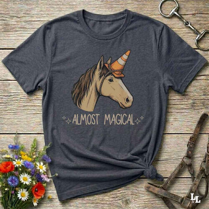 A dark heather gray graphic t-shirt laid flat on a rustic wooden surface. The shirt features a graphic of a white and brown horse wearing an orange and white striped cowboy horn, with the text 'ALMOST MAGICAL' printed below in a light beige font.