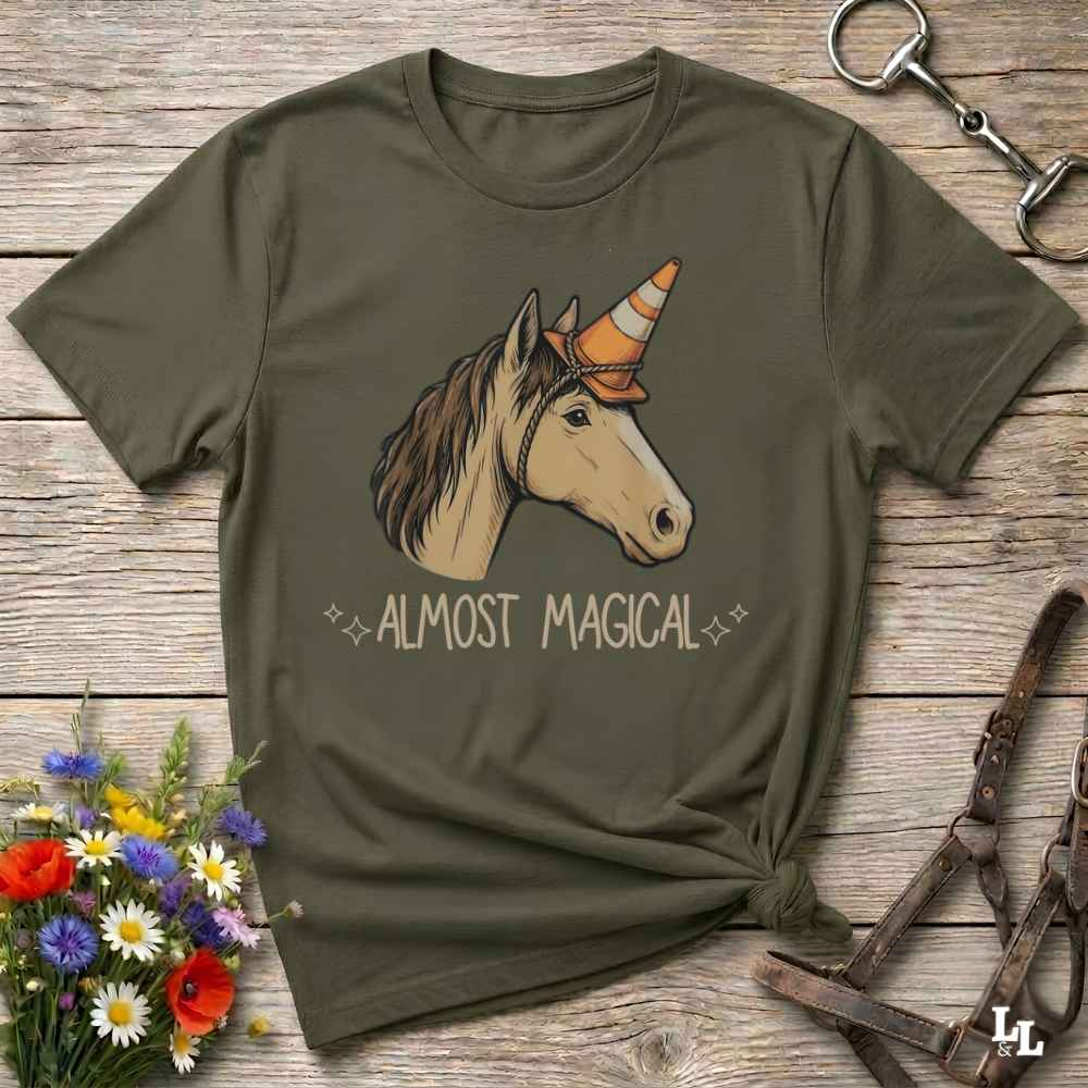 An olive green short-sleeve t-shirt with a graphic of a unicorn wearing an orange and white striped horn, above the text 'ALMOST MAGICAL'. The shirt is styled in a flat-lay on a rustic wooden background with a horse bridle, a horse bit, and a small bouquet of wildflowers.