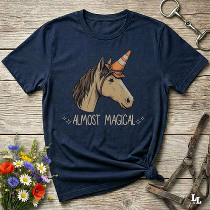 A navy blue short-sleeve t-shirt with a graphic of a unicorn wearing an orange and white striped horn, with the text 'ALMOST MAGICAL' printed below. The t-shirt is styled in a flat-lay on a rustic wooden background with equestrian-themed props like a halter, a horse bit, and a small bouquet of wildflowers.