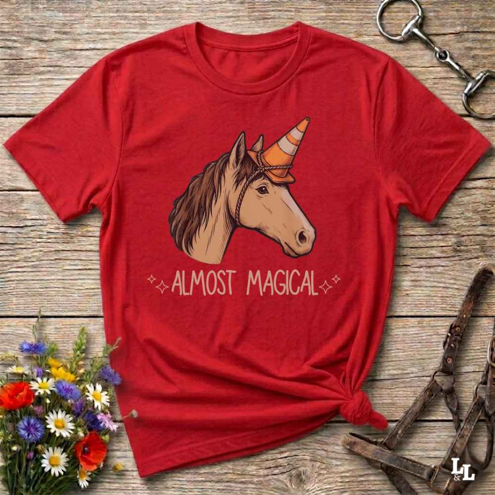 A red short-sleeve graphic T-shirt laid flat on a rustic wooden surface, featuring an illustrated unicorn head with a horn and the words 'ALMOST MAGICAL' below. The shirt is styled with flowers, a metal ring, and leather tack around it.