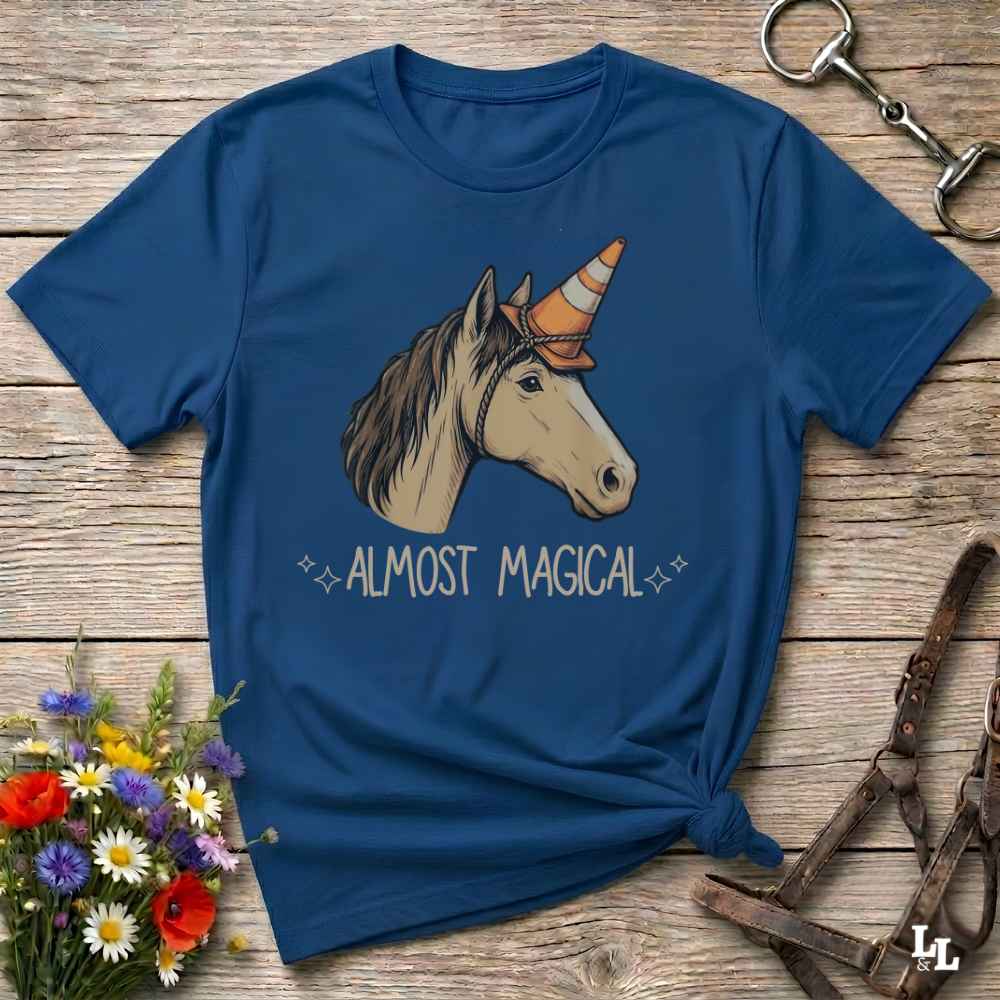 A navy blue graphic t-shirt laid flat on a rustic wooden surface, styled with a small bouquet of flowers and equestrian tack. The shirt features a graphic of a white horse with a striped orange and brown horn, above the text 'ALMOST MAGICAL'.