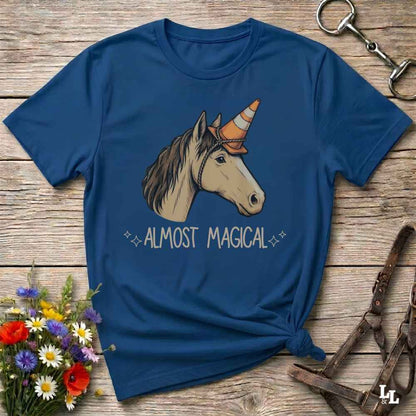 A navy blue graphic t-shirt laid flat on a rustic wooden surface, styled with a small bouquet of flowers and equestrian tack. The shirt features a graphic of a white horse with a striped orange and brown horn, above the text 'ALMOST MAGICAL'.