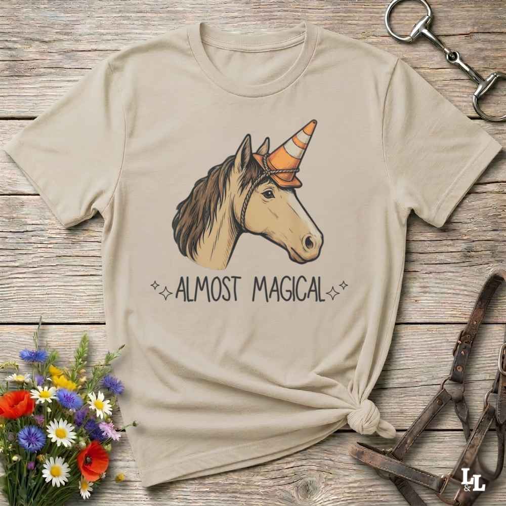 A beige graphic t-shirt featuring a unicorn with an orange party hat and the text 'ALMOST MAGICAL' underneath, styled in a flat-lay on a rustic wooden background with a flower bouquet and horse tack.
