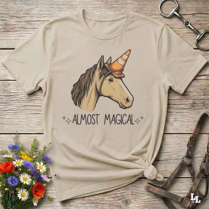 A beige graphic t-shirt featuring a unicorn with an orange party hat and the text 'ALMOST MAGICAL' underneath, styled in a flat-lay on a rustic wooden background with a flower bouquet and horse tack.