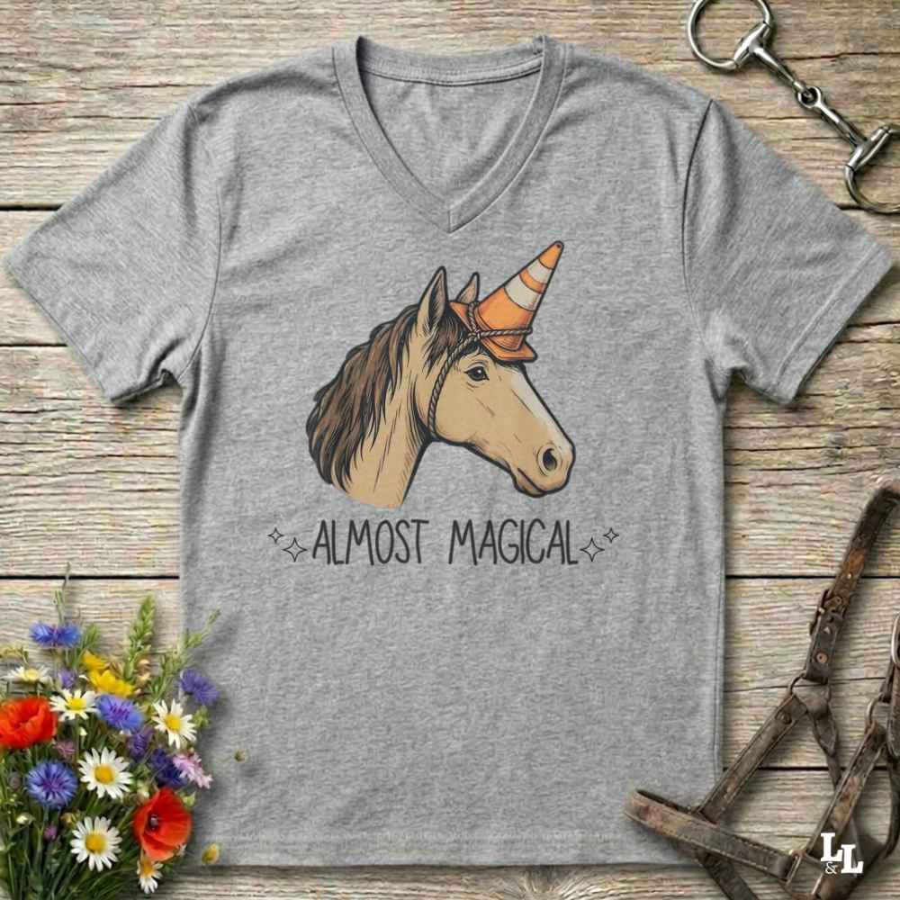 A heather gray V-neck t-shirt featuring a graphic of a horse head with a unicorn horn and the text 'ALMOST MAGICAL' underneath. The shirt is displayed flat on a rustic wooden background with equestrian and floral props.
