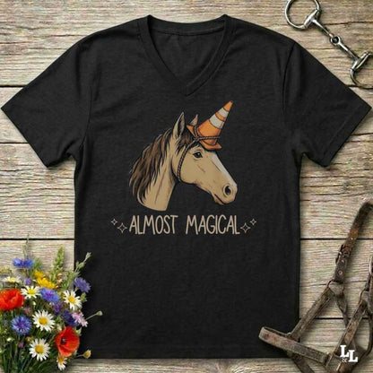 A black V-neck t-shirt laid flat on a rustic wooden surface, featuring a graphic of a horse head with a traffic cone horn and the text 'ALMOST MAGICAL'. The shirt is styled with equestrian accessories (bridle, horse bit) and a small bouquet of wildflowers.