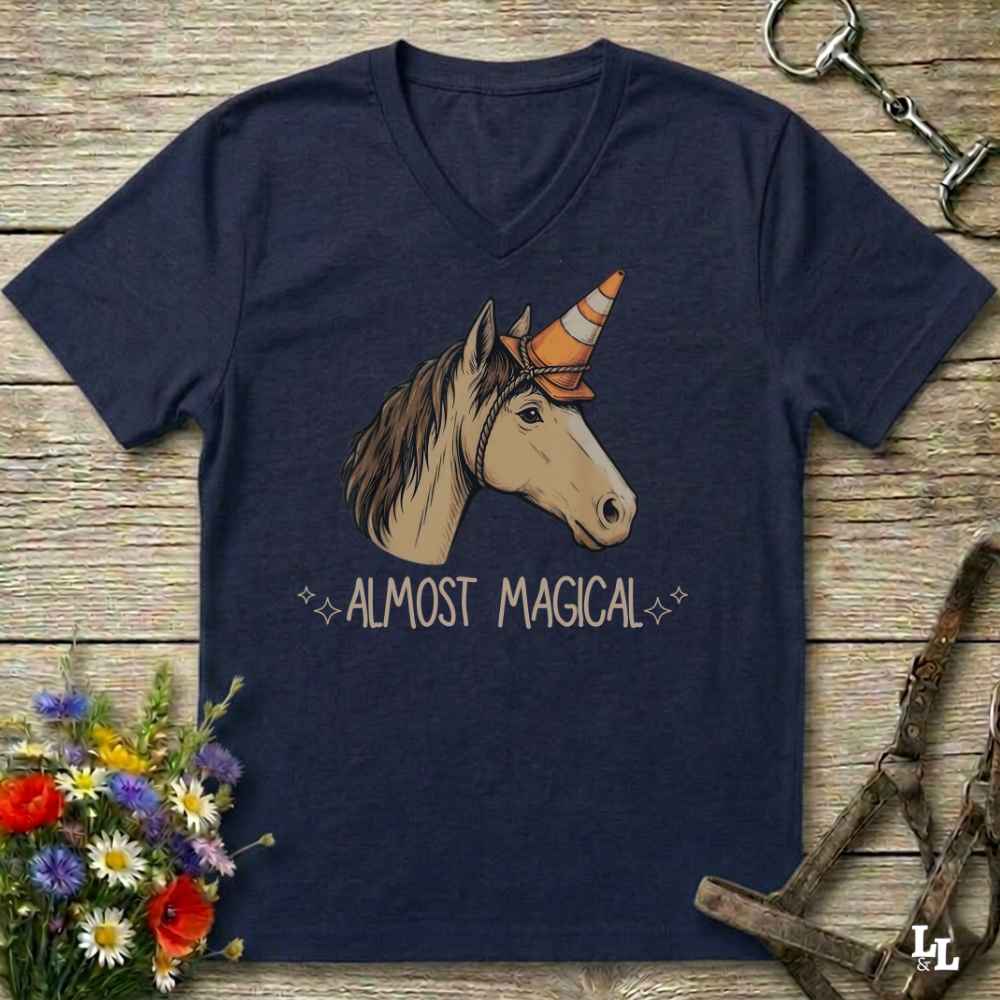 A navy blue V-neck t-shirt laid flat on a rustic wooden surface, featuring a graphic of a horse head with a unicorn horn and the text 'ALMOST MAGICAL'. The shirt is styled with equestrian tack, a bouquet of wildflowers, and a metal bit.