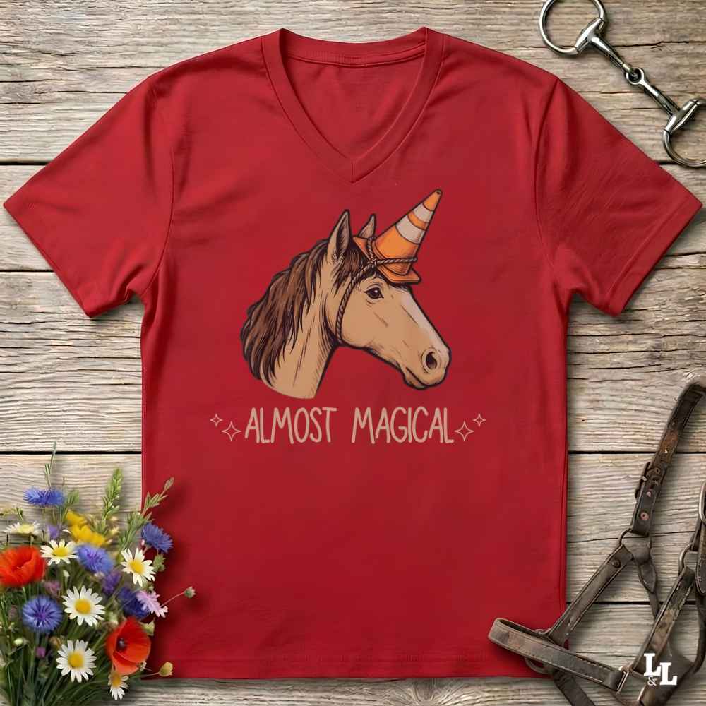 A red V-neck t-shirt laid flat on a rustic wooden surface, featuring a graphic of a brown horse with a unicorn horn and the text 'ALMOST MAGICAL' in white. The shirt is styled with equestrian accessories (a horse bit and halter) and a small bouquet of wildflowers.