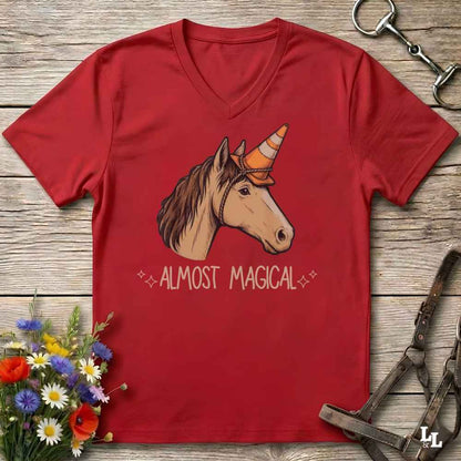 A red V-neck t-shirt laid flat on a rustic wooden surface, featuring a graphic of a brown horse with a unicorn horn and the text 'ALMOST MAGICAL' in white. The shirt is styled with equestrian accessories (a horse bit and halter) and a small bouquet of wildflowers.