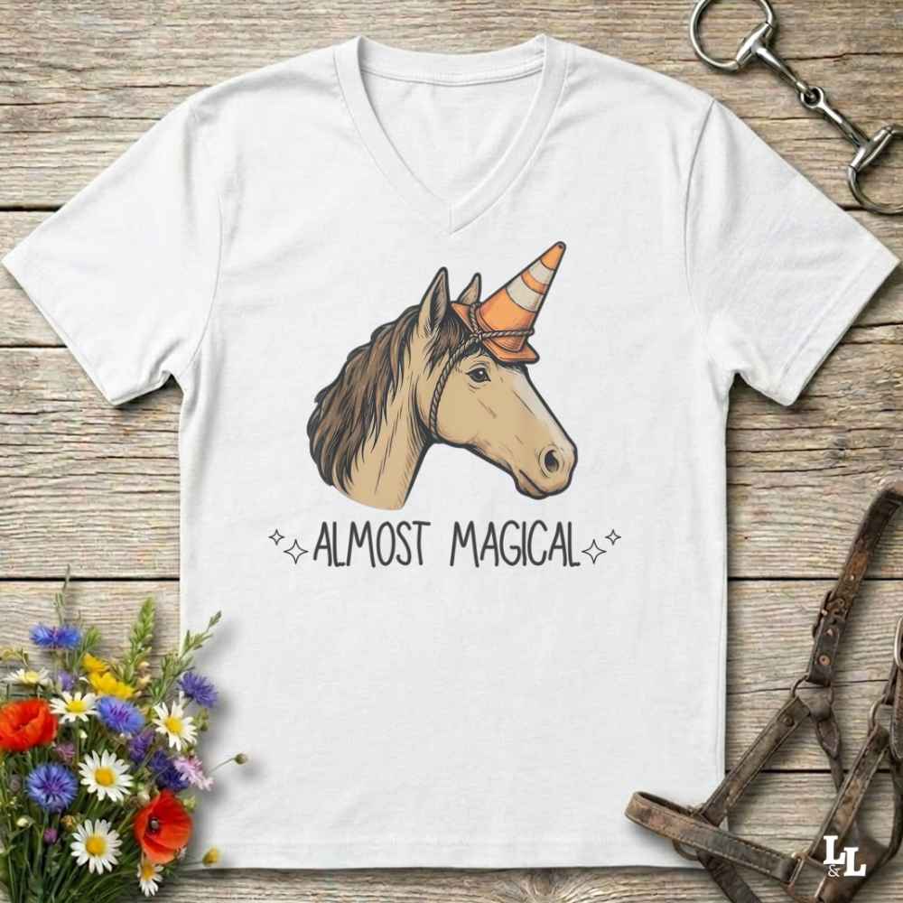 A white V-neck t-shirt laid flat on a rustic wooden surface, featuring a graphic of a horse head with an orange-and-white striped unicorn horn and the text 'ALMOST MAGICAL'. The shirt is styled with flowers, a horse bit, and a leather bridle.
