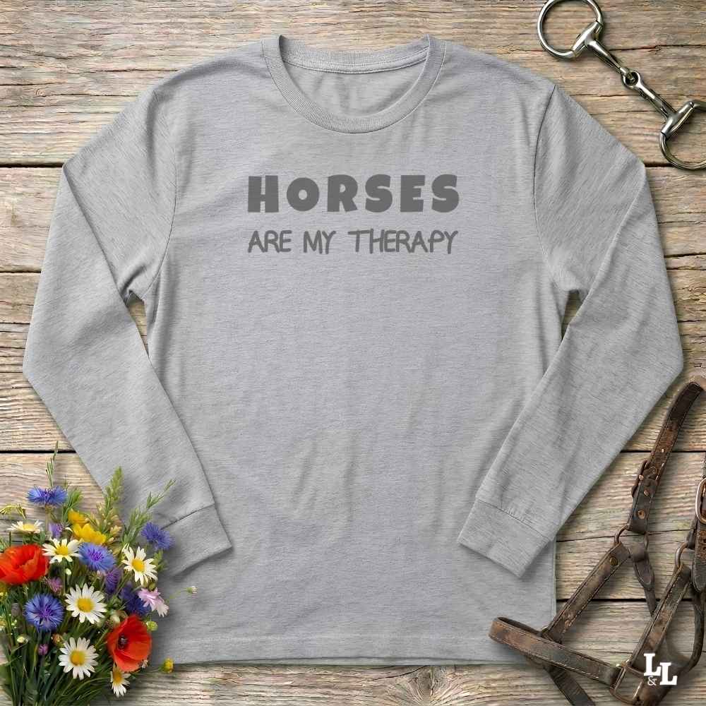 A light gray long-sleeve t-shirt with the text 'HORSES ARE MY THERAPY' printed in dark gray on the chest, laid flat on a rustic wooden background with decorative equestrian and floral props.