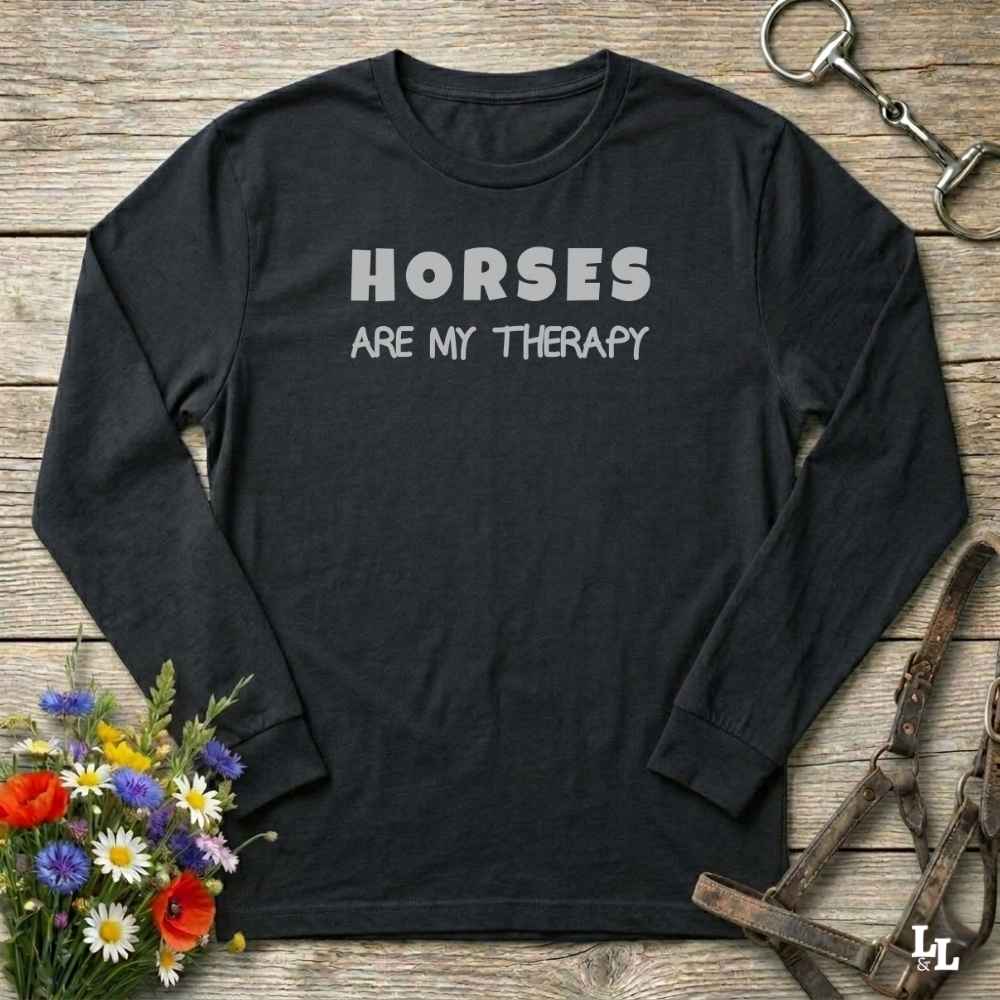 A black long-sleeve t-shirt with the text 'HORSES ARE MY THERAPY' printed in white on the chest, laid flat on a rustic wooden surface with equestrian and floral props.