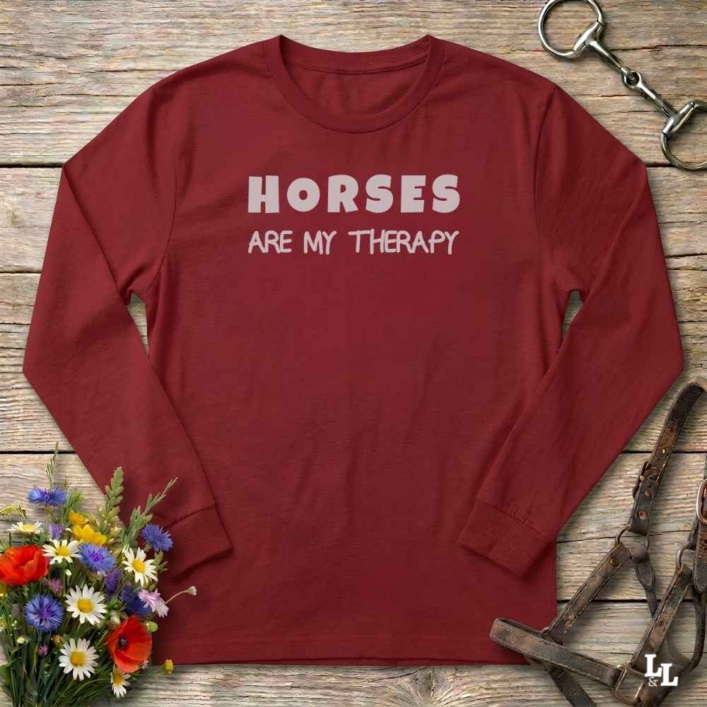 A long-sleeve crewneck shirt in burgundy red with the text 'HORSES ARE MY THERAPY' printed in white across the chest. The shirt is laid flat on a rustic wooden surface, surrounded by equestrian-themed props including a horse bit, leather bridle, and a small bouquet of colorful flowers.