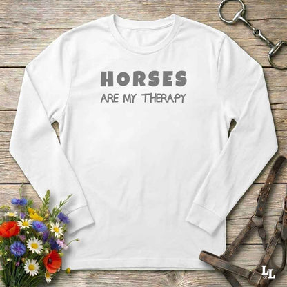 A white long-sleeve t-shirt with the text 'HORSES ARE MY THERAPY' printed in gray on the chest, laid flat on a rustic wooden background with equestrian and floral props.