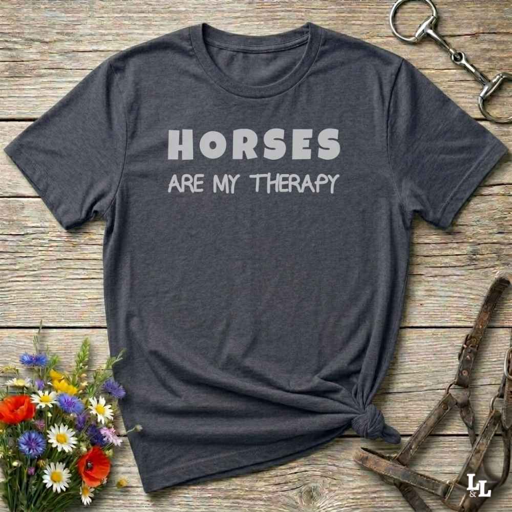 A dark heather gray t-shirt with the text 'HORSES ARE MY THERAPY' printed in white across the chest, styled in a flat-lay on a rustic wooden background with equestrian accessories and flowers.