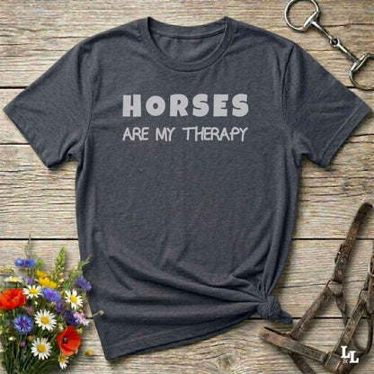 A dark heather gray t-shirt with the text 'HORSES ARE MY THERAPY' printed in white across the chest, styled in a flat-lay on a rustic wooden background with equestrian accessories and flowers.