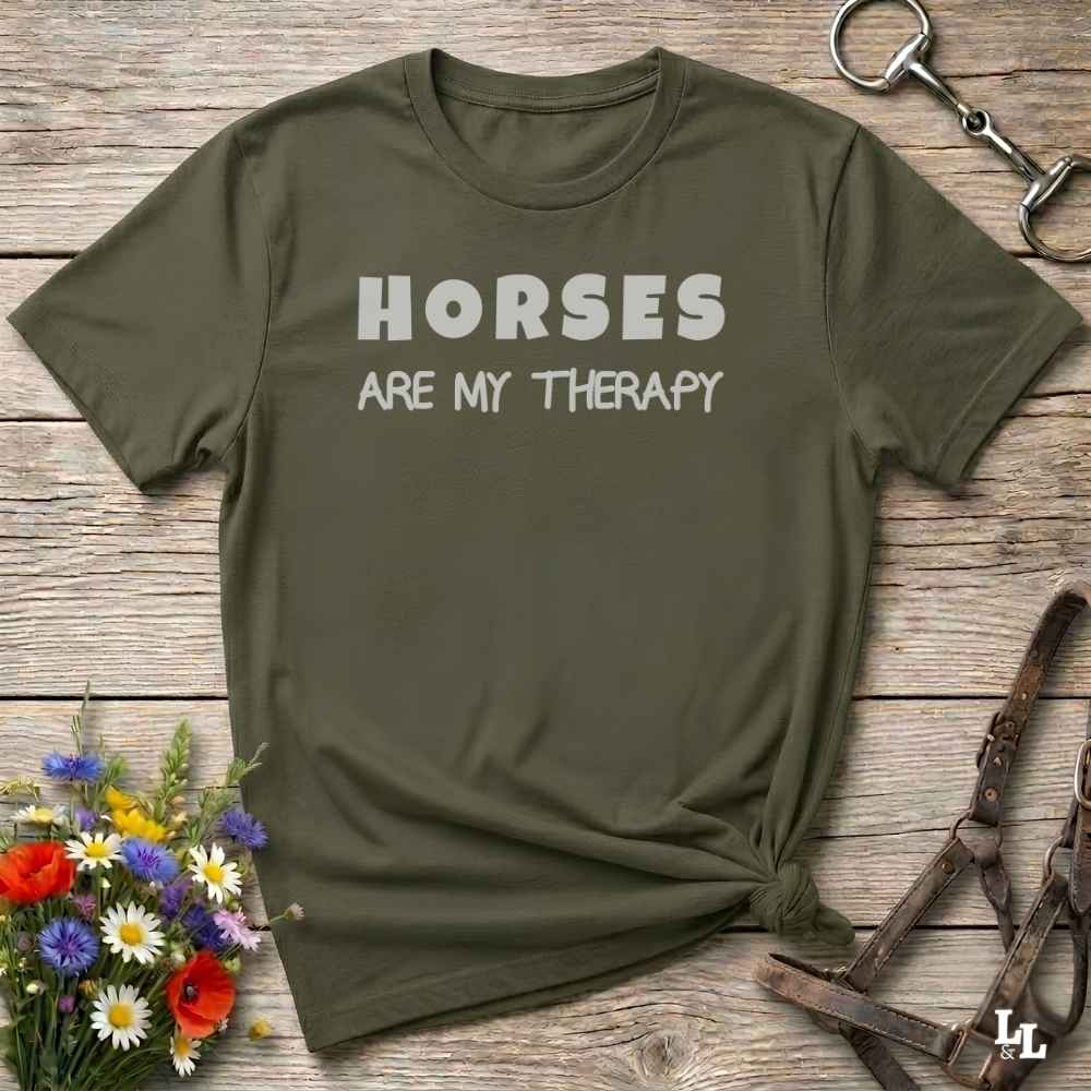 An olive green crewneck t-shirt with the text 'HORSES ARE MY THERAPY' printed in white across the chest, styled in a flat-lay on a rustic wooden background with equestrian accessories and wildflowers.