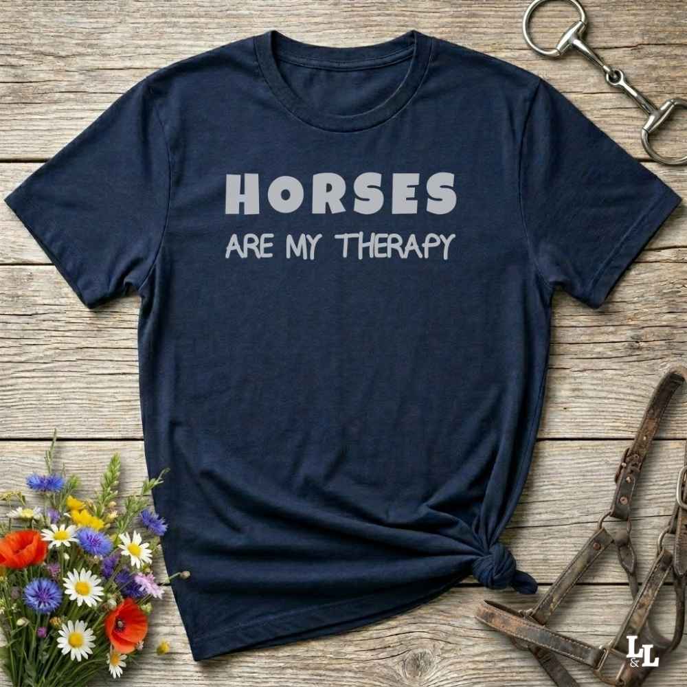 A navy blue short-sleeve graphic t-shirt with the text 'HORSES ARE MY THERAPY' printed in white on the front. The shirt is styled in a flat-lay arrangement on a rustic wooden background, surrounded by decorative elements including a small bouquet of colorful wildflowers, a metal horse bit, and a brown leather halter.