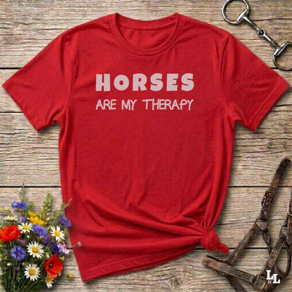 A red short-sleeve crewneck t-shirt with the text 'HORSES ARE MY THERAPY' printed in white across the chest, styled in a flat lay on a rustic wooden background with equestrian accessories and wildflowers.
