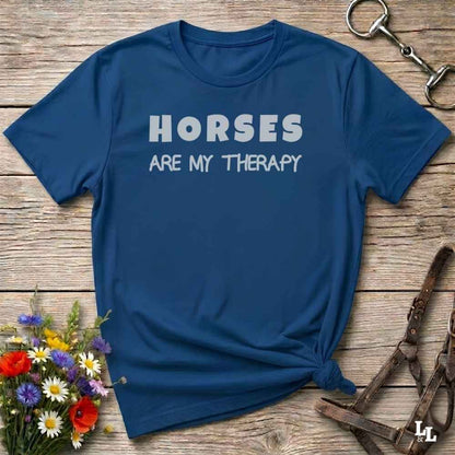 A navy blue short-sleeve t-shirt with the text 'HORSES ARE MY THERAPY' printed in white across the chest, styled in a flat lay on a rustic wooden surface with equestrian and floral props.