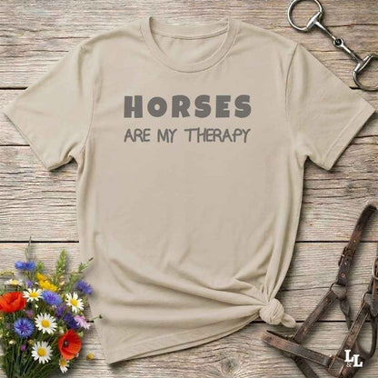 A beige graphic t-shirt with the text 'HORSES ARE MY THERAPY' printed in gray, styled in a flat-lay on a rustic wooden background with equestrian accessories and wildflowers.