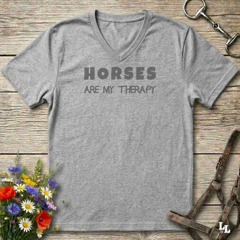 A heather gray V-neck t-shirt with the text 'HORSES ARE MY THERAPY' printed in dark gray on the chest, laid flat on a rustic wooden surface with equestrian-themed props like a horse bit, halter, and wildflowers.
