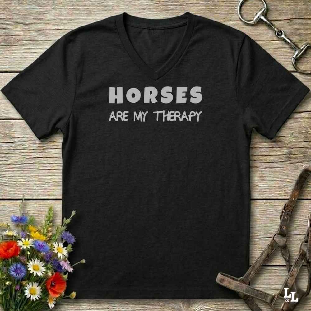 A black V-neck t-shirt with the text 'HORSES ARE MY THERAPY' printed in white across the chest, displayed on a rustic wooden background with equestrian and floral props.