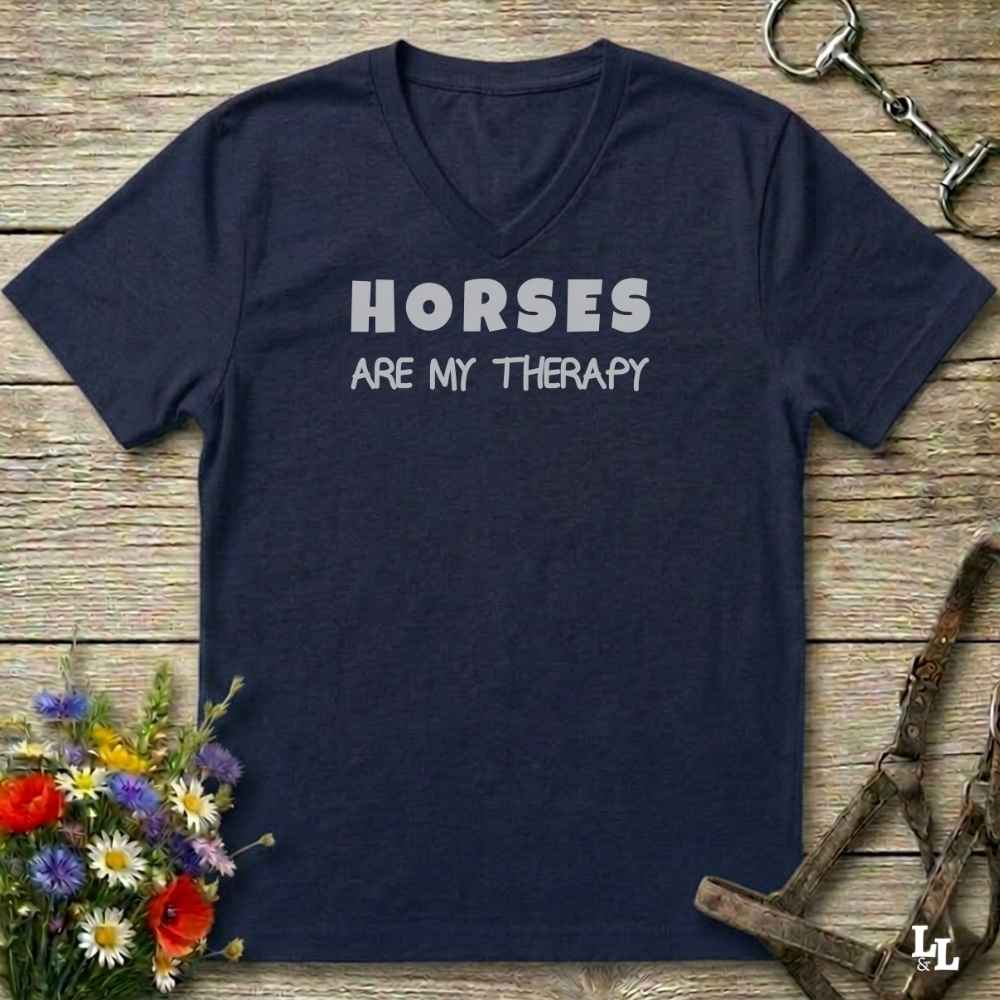 A navy blue V-neck t-shirt with the text 'HORSES ARE MY THERAPY' printed in white across the chest, displayed on a rustic wooden background with equestrian-themed props like a halter, bit, and wildflowers.