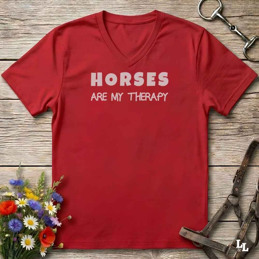 A red V-neck t-shirt with the text 'HORSES ARE MY THERAPY' printed in white across the chest, laid flat on a rustic wooden surface with decorative equestrian and floral props.