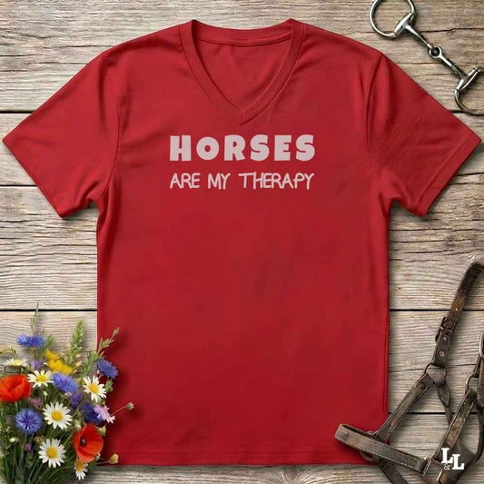 A red V-neck t-shirt with the text 'HORSES ARE MY THERAPY' printed in white across the chest, laid flat on a rustic wooden surface with decorative equestrian and floral props.