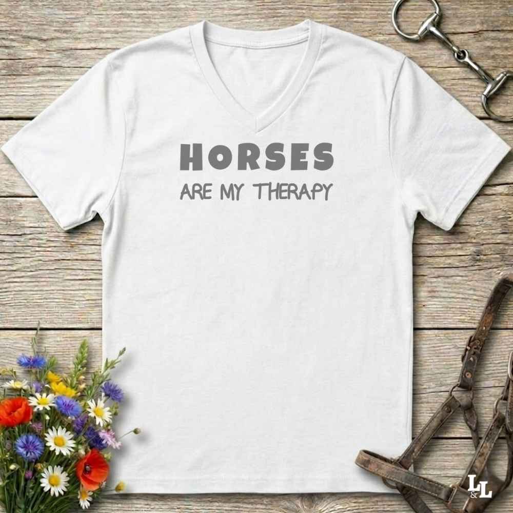 A white V-neck t-shirt with the text 'HORSES ARE MY THERAPY' printed in gray across the chest, displayed on a rustic wooden background with equestrian and floral props.