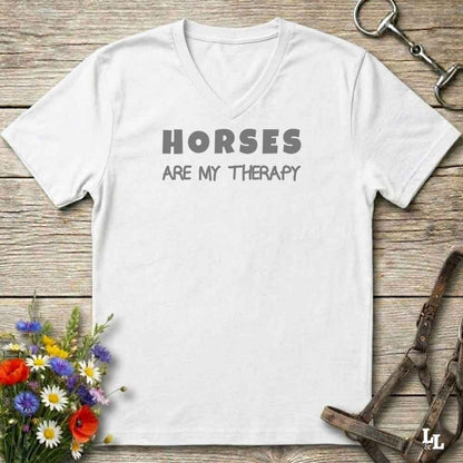 A white V-neck t-shirt with the text 'HORSES ARE MY THERAPY' printed in gray across the chest, displayed on a rustic wooden background with equestrian and floral props.