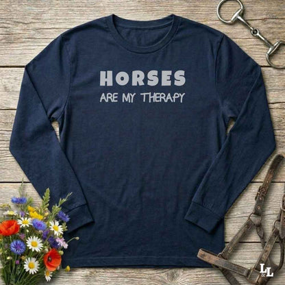 A navy blue long-sleeve t-shirt with the text 'HORSES ARE MY THERAPY' printed in white on the chest, styled in a flat-lay on a rustic wooden background with equestrian and floral props.