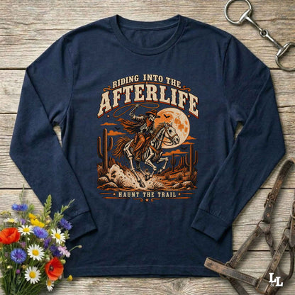 A navy blue long-sleeve t-shirt with a western-themed graphic print, laid flat on a rustic wooden background with decorative props including flowers, a horse bit, and a leather bridle.