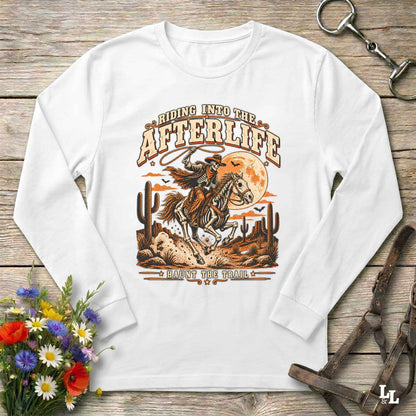 A white long-sleeve t-shirt with a western-themed graphic print, laid flat on a rustic wooden background with decorative props including flowers, a horse bit, and a leather bridle.