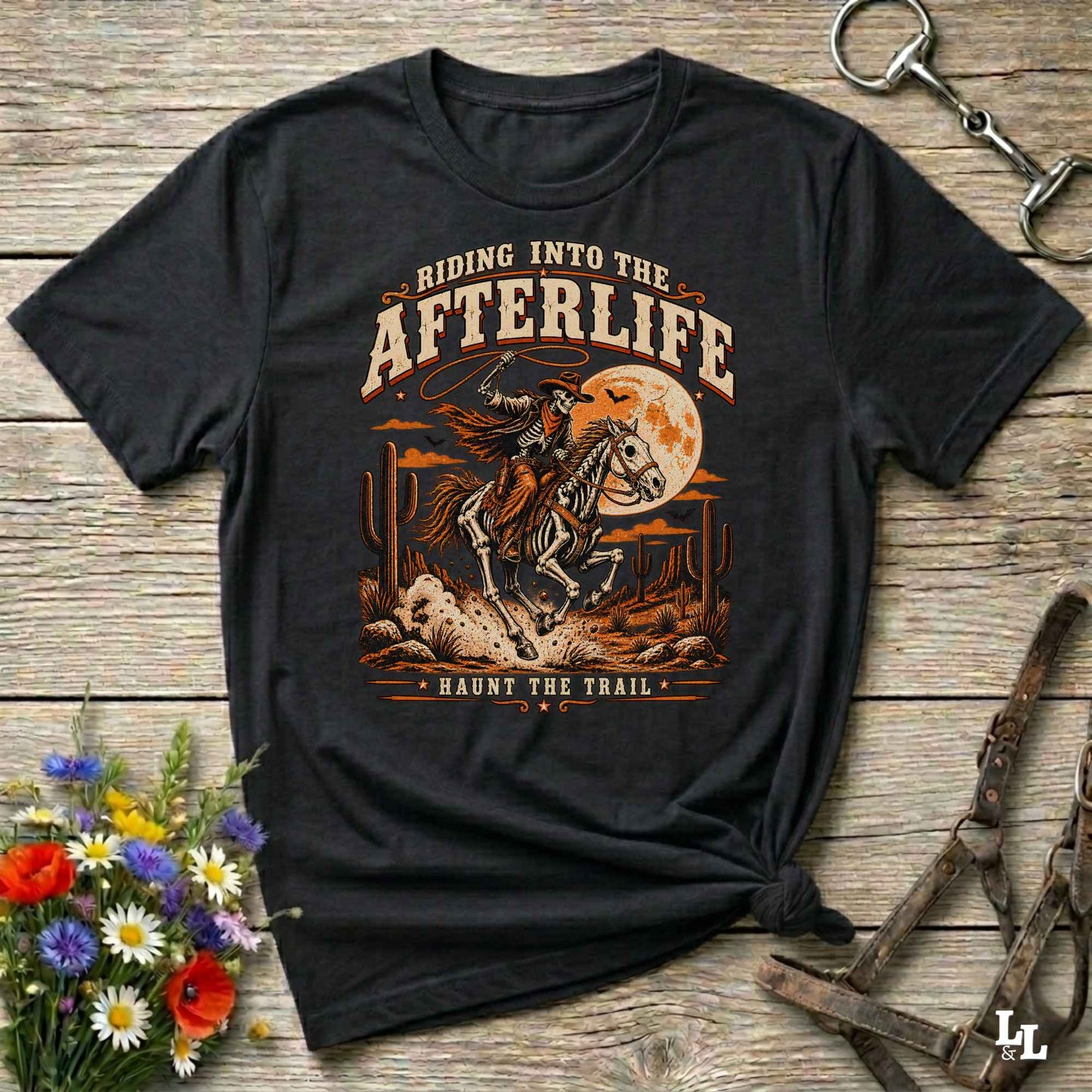 A black graphic t-shirt featuring a western-style design of a skeleton cowboy riding a horse in front of a full moon, with the text 'RIDING INTO THE AFTERLIFE' and 'HAUNTS THE TRAIL'. The shirt is displayed on a rustic wooden background with equestrian accessories and flowers.