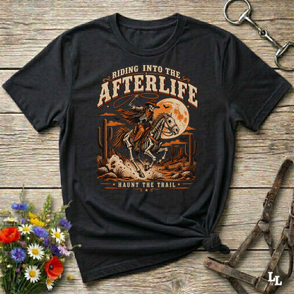 A black graphic t-shirt featuring a western-style design of a skeleton cowboy riding a horse in front of a full moon, with the text 'RIDING INTO THE AFTERLIFE' and 'HAUNTS THE TRAIL'. The shirt is displayed on a rustic wooden background with equestrian accessories and flowers.