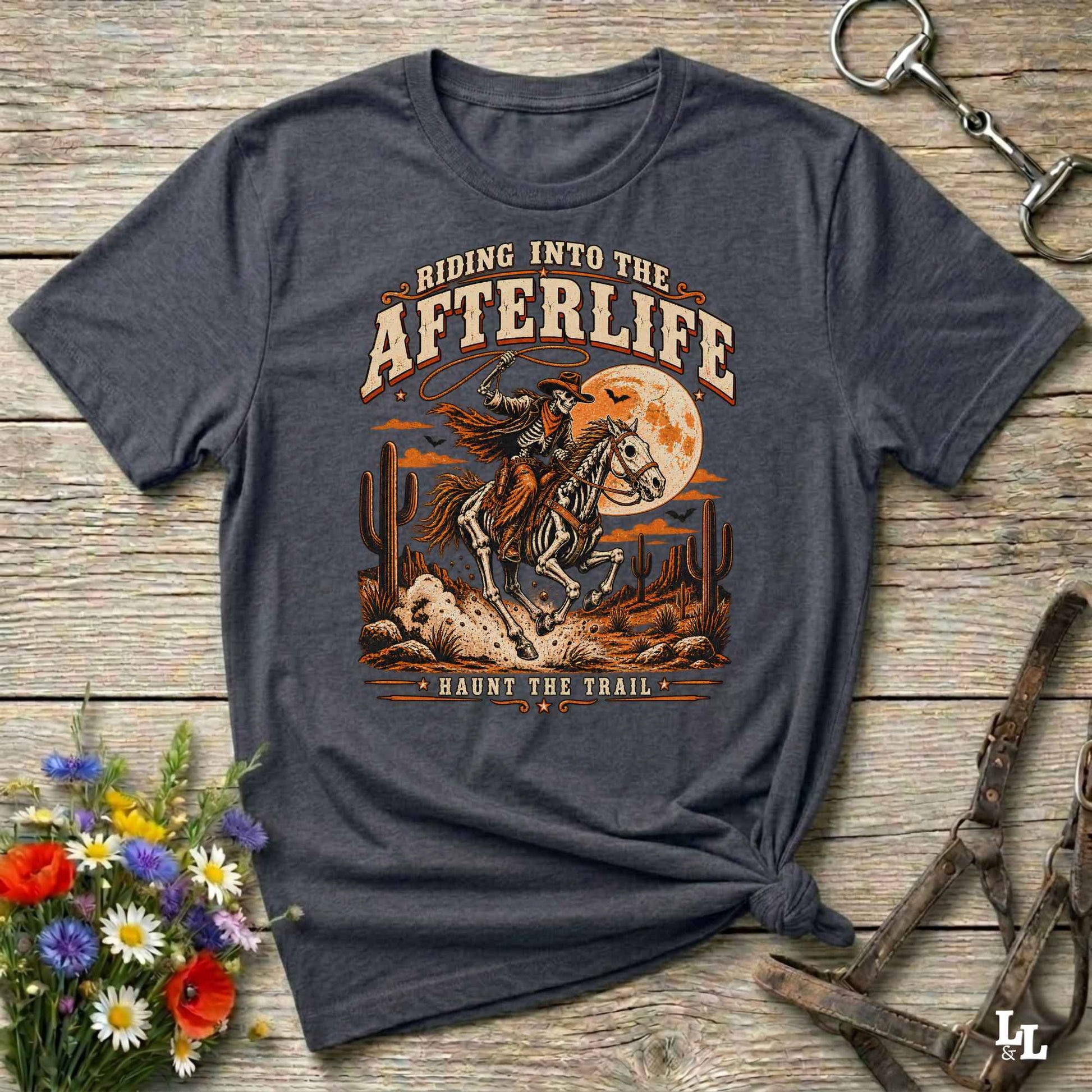 A dark heather gray graphic t-shirt featuring a western-themed design with a cowboy on a bucking horse in front of a full moon, laid flat on a rustic wooden background with horse bits and flowers as props.