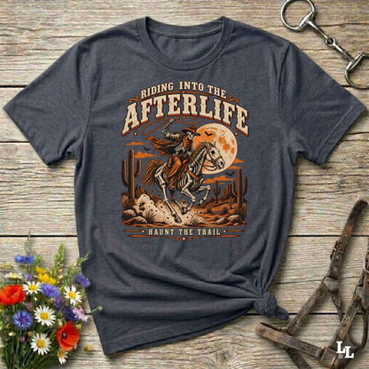 A dark heather gray graphic t-shirt featuring a western-themed design with a cowboy on a bucking horse in front of a full moon, laid flat on a rustic wooden background with horse bits and flowers as props.