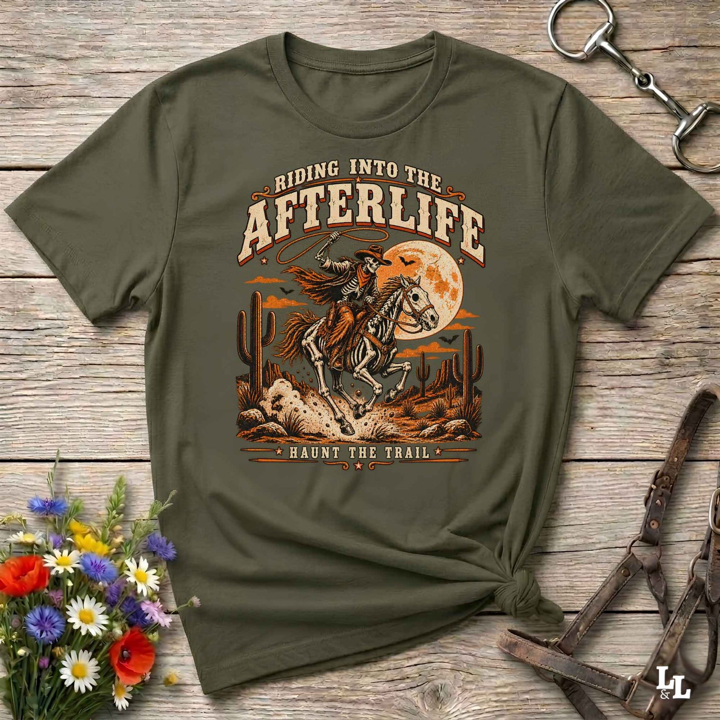 An olive green graphic t-shirt featuring a western design with a cowboy on a galloping horse in front of a full moon, with the text 'RIDING INTO THE AFTERLIFE' and 'HAUNT THE TRAIL'. The t-shirt is styled in a flat-lay on a rustic wooden background with horse tack, flowers, and a leather halter.