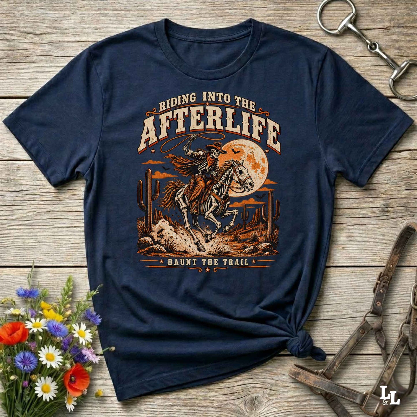 A navy blue graphic t-shirt laid flat on a rustic wooden surface, styled with a small bouquet of wildflowers, a horse bit, and a leather bridle. The t-shirt features a detailed western-themed graphic of a skeleton cowboy riding a horse under a full moon with the text 'RIDING INTO THE AFTERLIFE' and 'HAUNT THE TRAIL'.