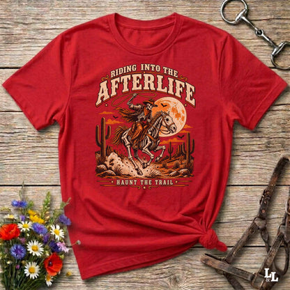 A red short-sleeve graphic T-shirt laid flat on a rustic wooden surface, styled with flowers, a horse bit, and a leather bridle. The shirt features a large western-style print of a rider on a horse in front of a full moon, with the text 'Riding Into the AFTERLIFE' and 'HAUNT THE TRAIL'.